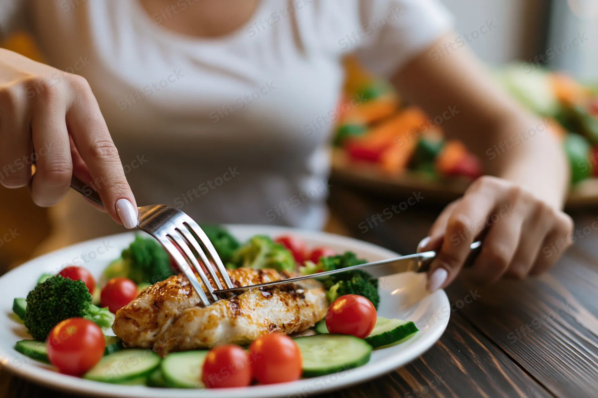 Woman cutting grilled chicken breast with vegetables on a white plate at a wooden table