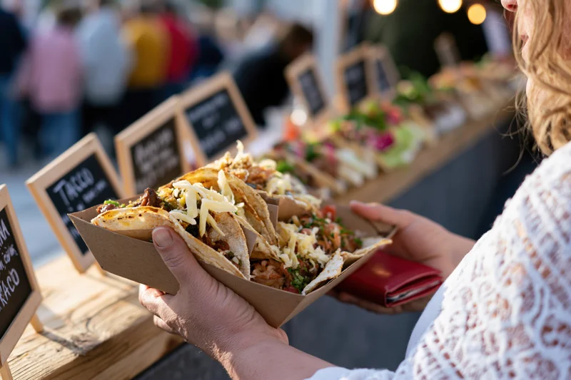 Woman holding a paper tray of loaded street tacos at an outdoor food market