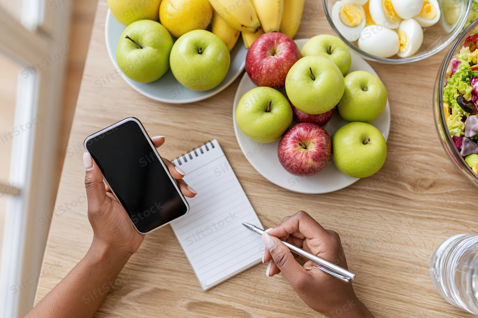Woman’s hands writing in a spiral notepad while holding a smartphone on a kitchen table with fruit and breakfast items