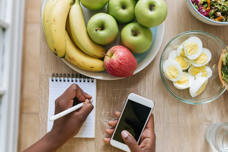 Woman's hands writing in a spiral notepad while holding a smartphone over a breakfast table with fruit, eggs and salad