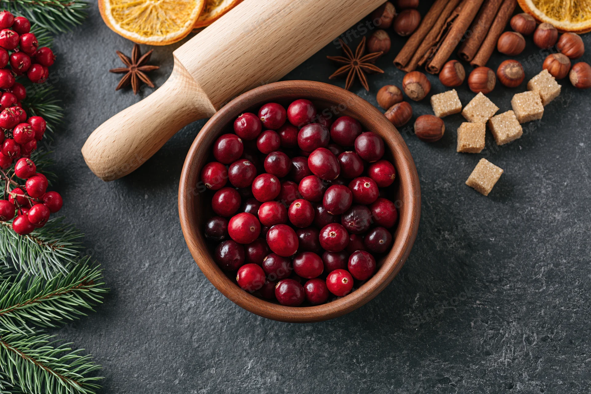 Wooden bowl filled with fresh cranberries surrounded by holiday spices and evergreen sprigs on dark surface