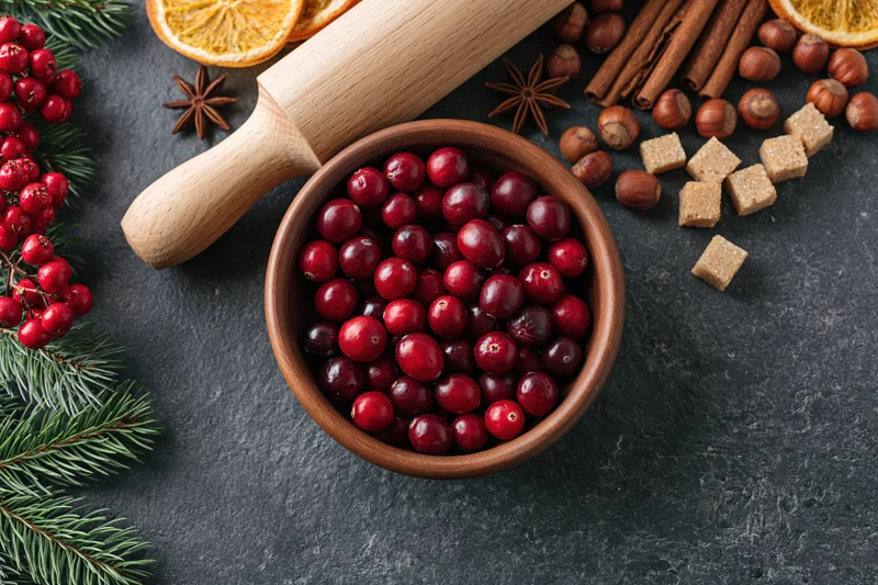 Wooden bowl filled with fresh cranberries surrounded by holiday spices and evergreen sprigs on dark surface