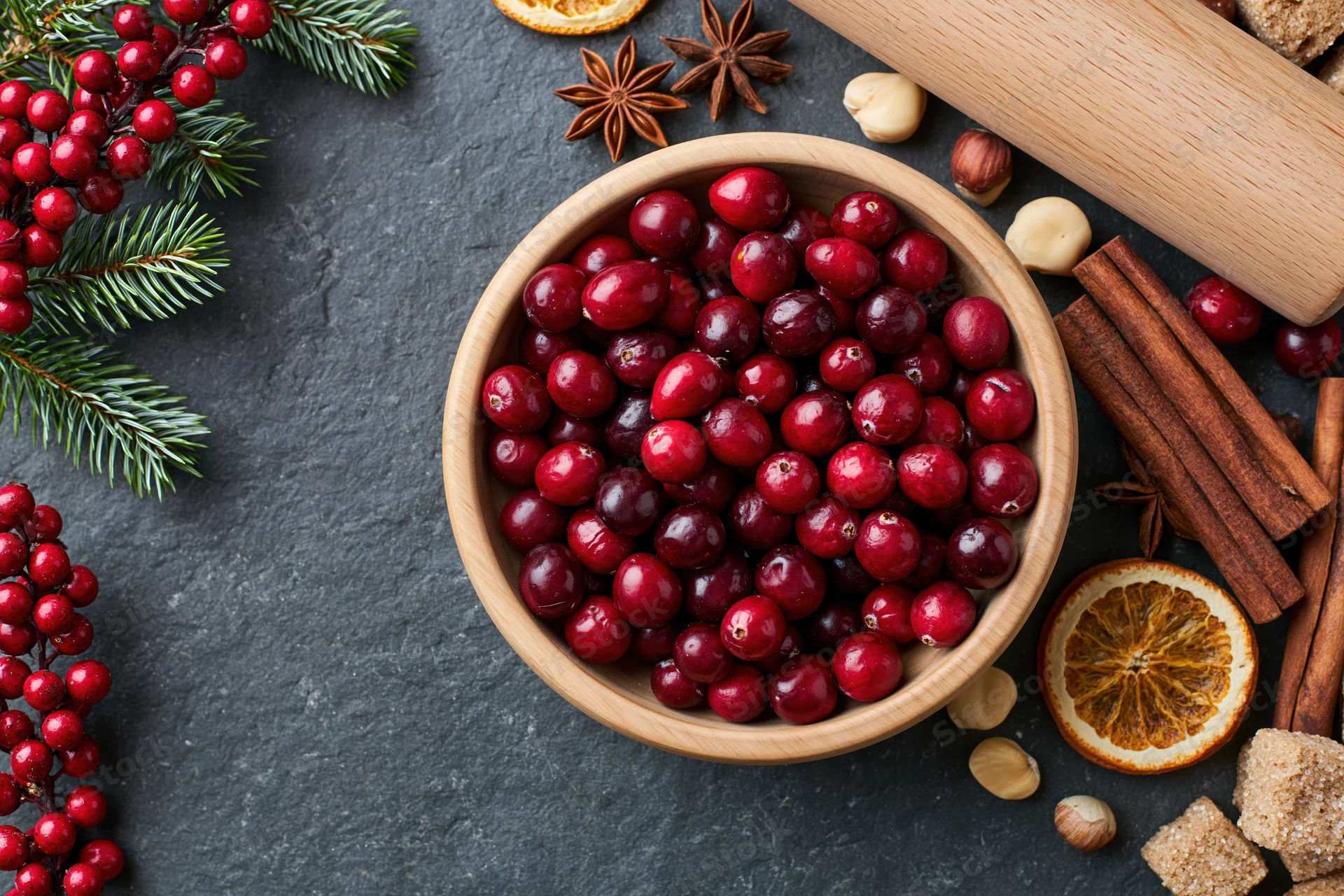 Wooden bowl filled with fresh cranberries surrounded by holiday decorations and baking ingredients on dark slate surface