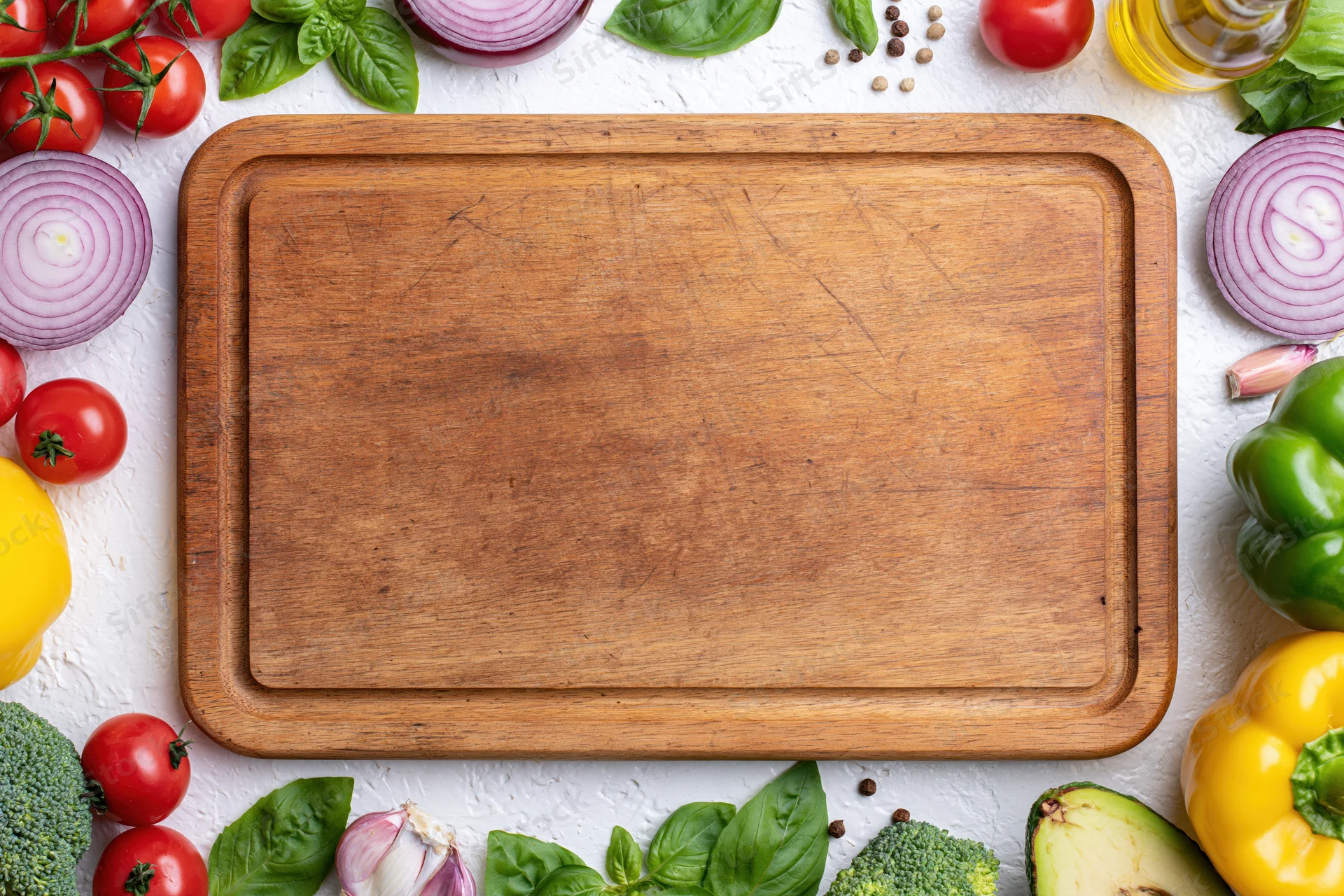 Worn wooden cutting board surrounded by fresh vegetables and herbs on white surface