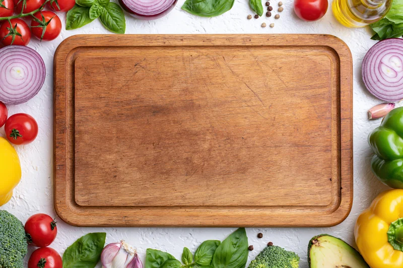 Worn wooden cutting board surrounded by fresh vegetables and herbs on white surface