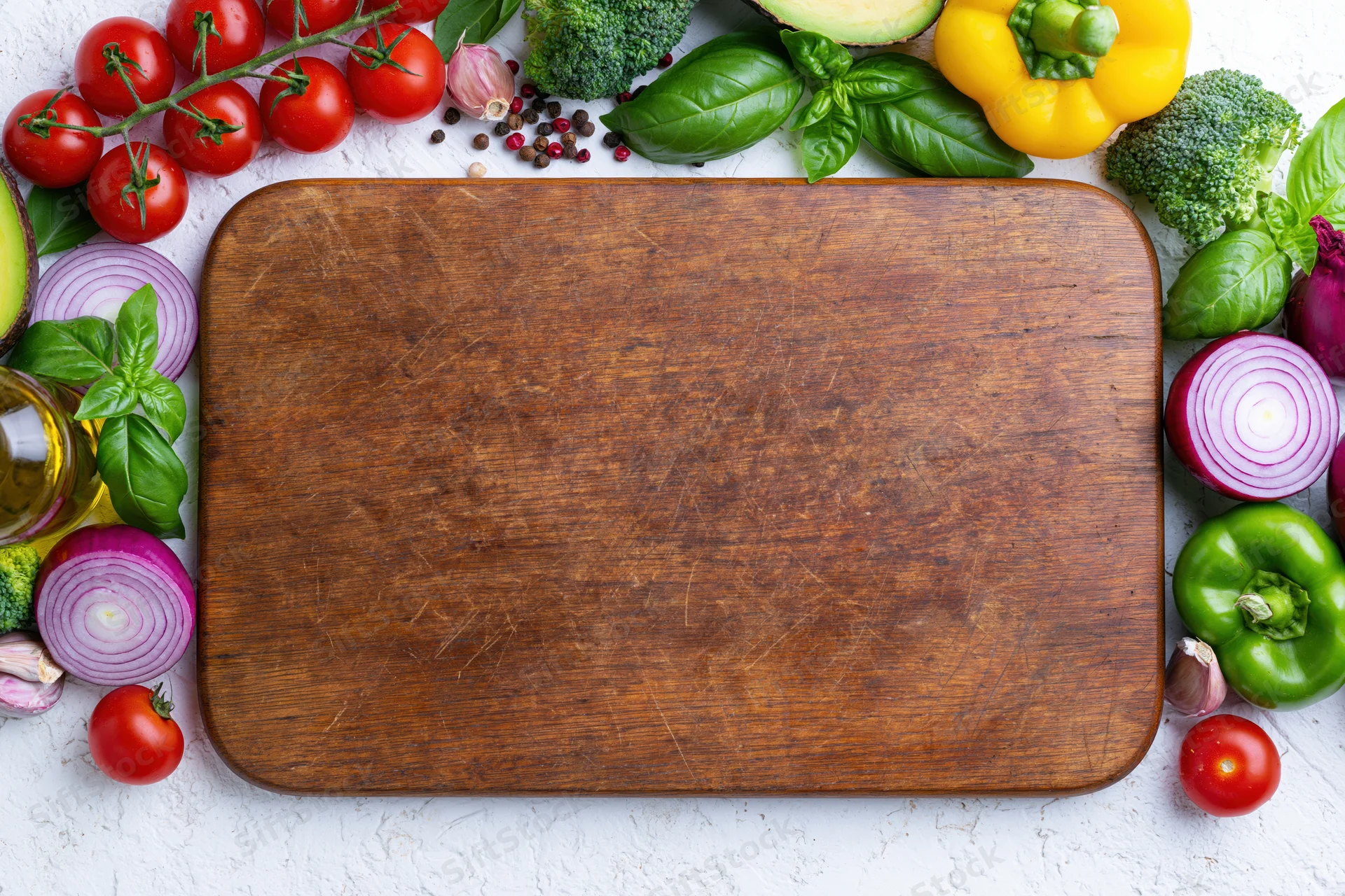 Worn wooden cutting board surrounded by fresh vegetables and herbs on white countertop