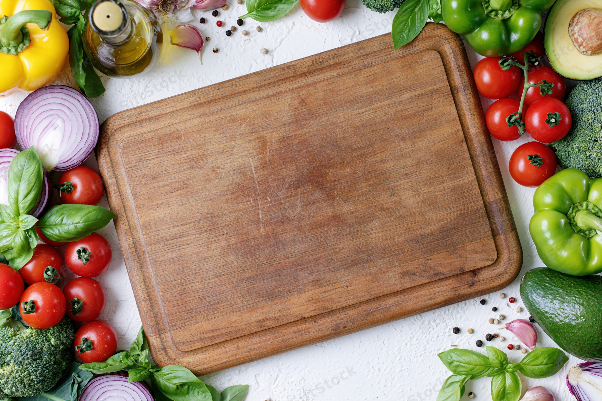 Worn wooden cutting board surrounded by fresh vegetables and herbs on white surface