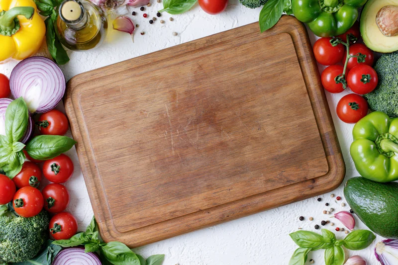 Worn wooden cutting board surrounded by fresh vegetables and herbs on white surface