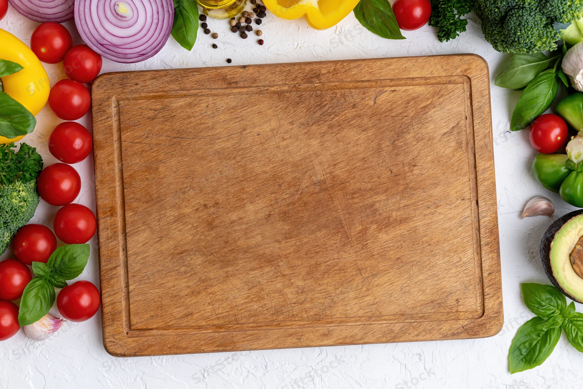 Worn wooden cutting board centered on white surface surrounded by fresh vegetables and herbs