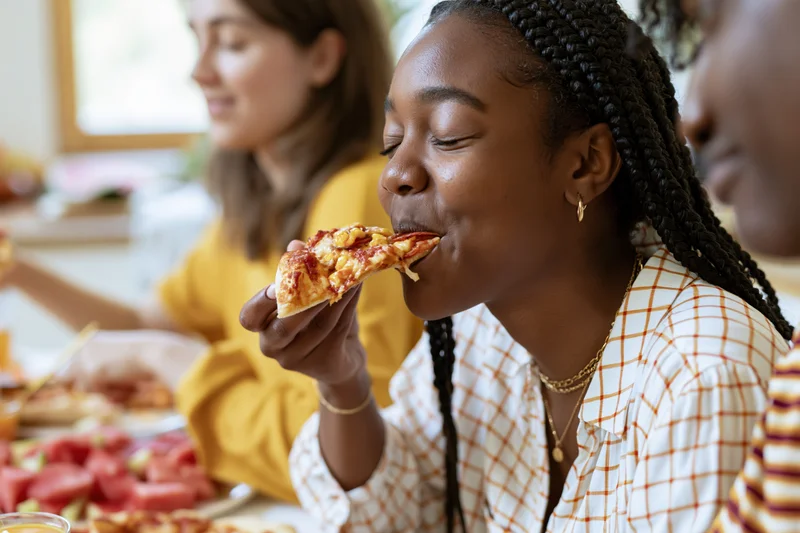 Young black woman eating pizza slice at dining table with friends