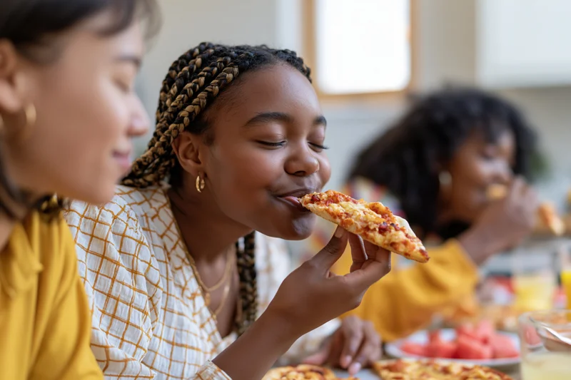 Young black woman with box braids eating pizza at dining table with friends