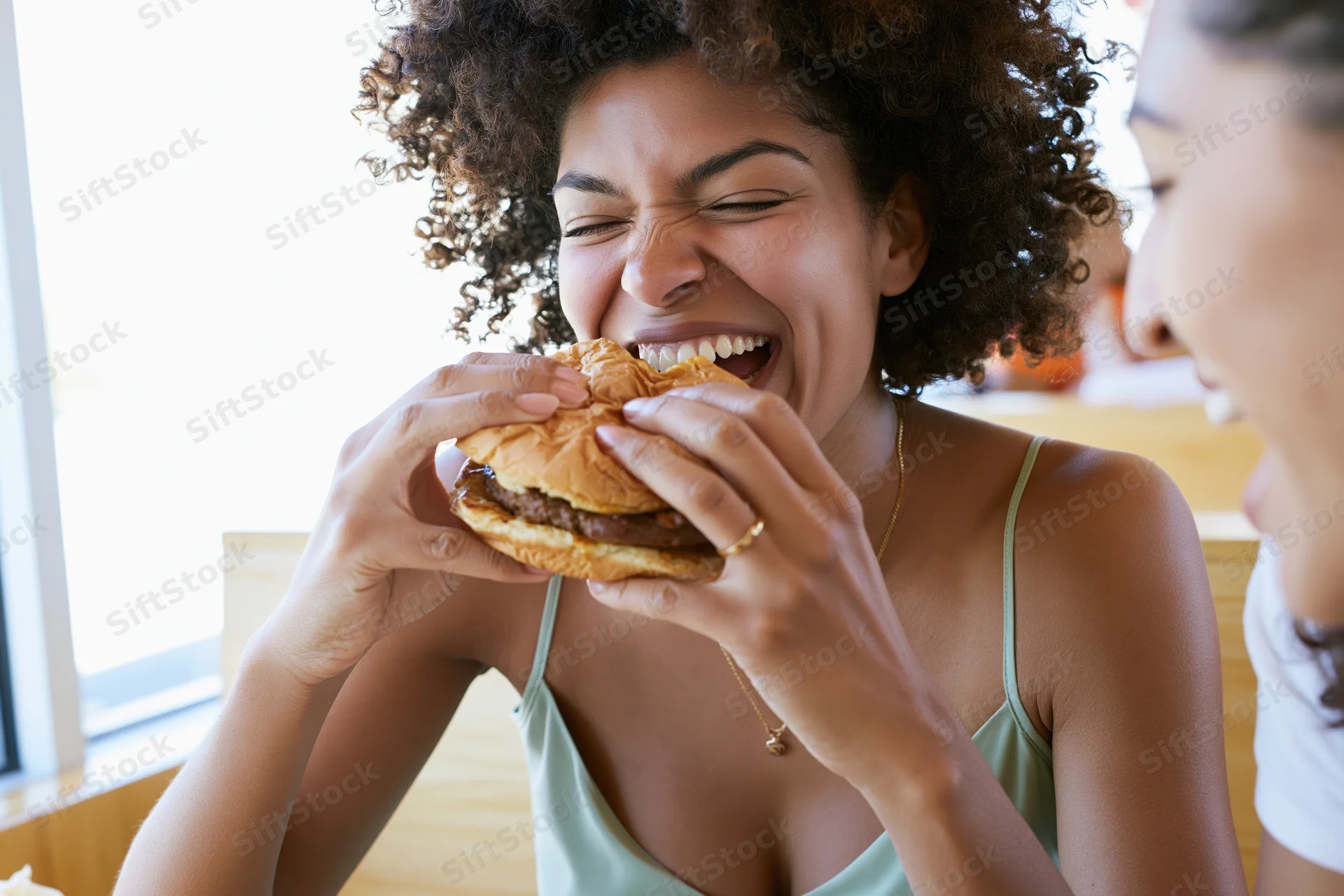 Young black woman laughing mid-bite while eating a burger in a restaurant with a friend
