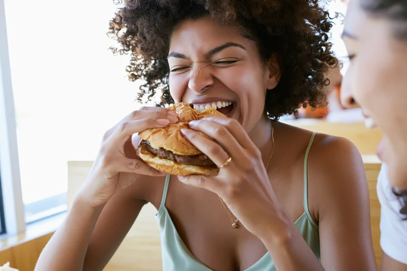 Young black woman laughing mid-bite while eating a burger in a restaurant with a friend
