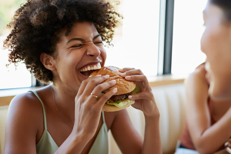 Young black woman laughing while eating a burger in a restaurant booth with a friend