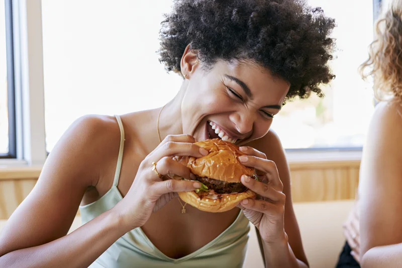 Young black woman laughing while taking a big bite of a burger in a restaurant