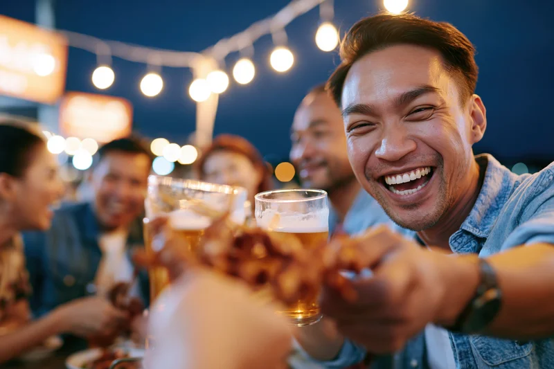 Young man smiling broadly while toasting with beer at outdoor night party with friends