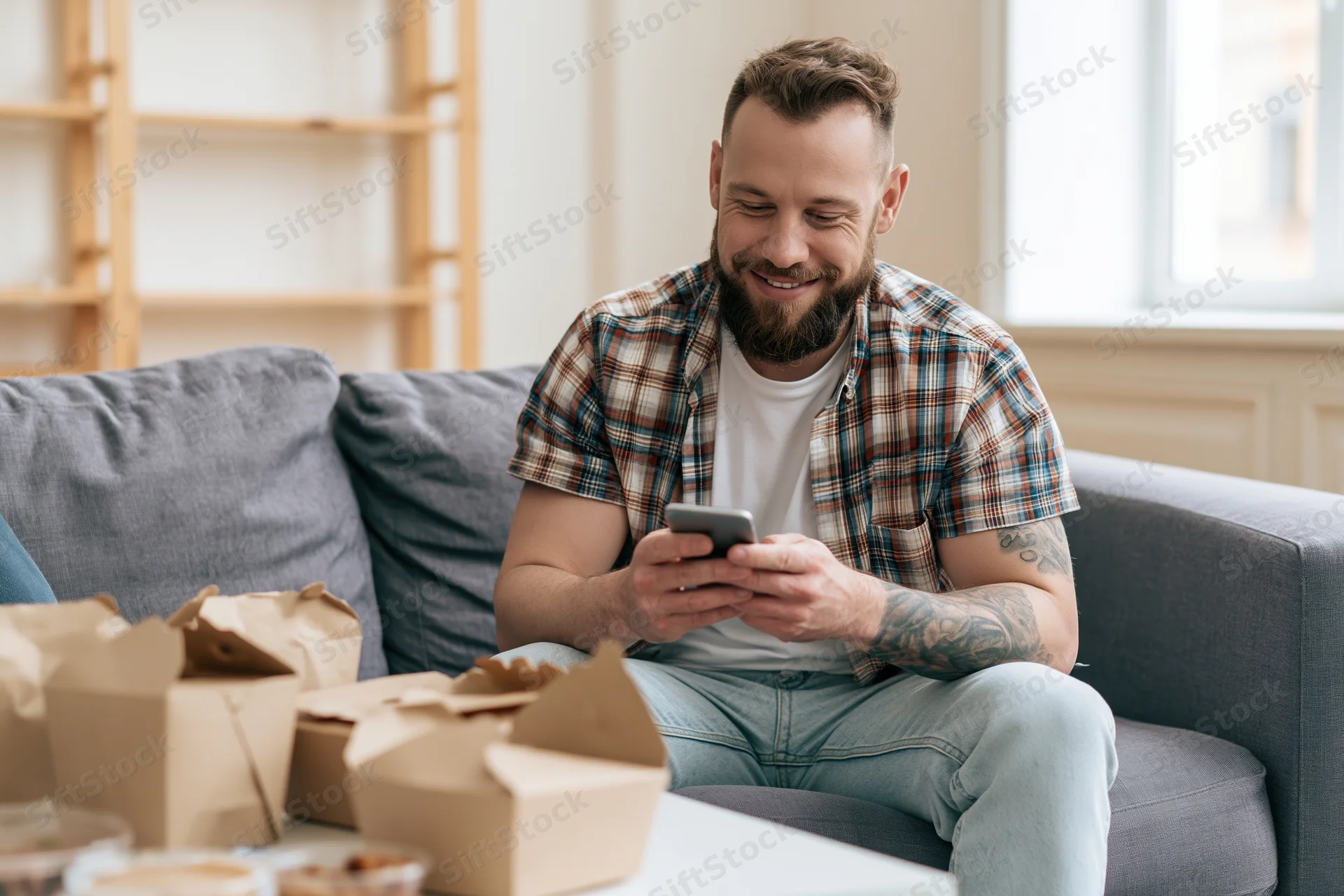 Man smiling while using smartphone on grey sofa with takeout boxes nearby