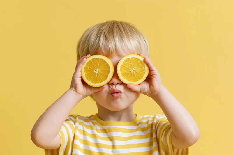Young blonde toddler holding orange halves over eyes in front of yellow background