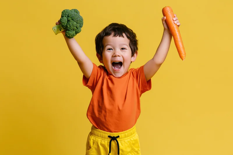 Young boy laughing with arms raised holding broccoli and carrot against yellow background