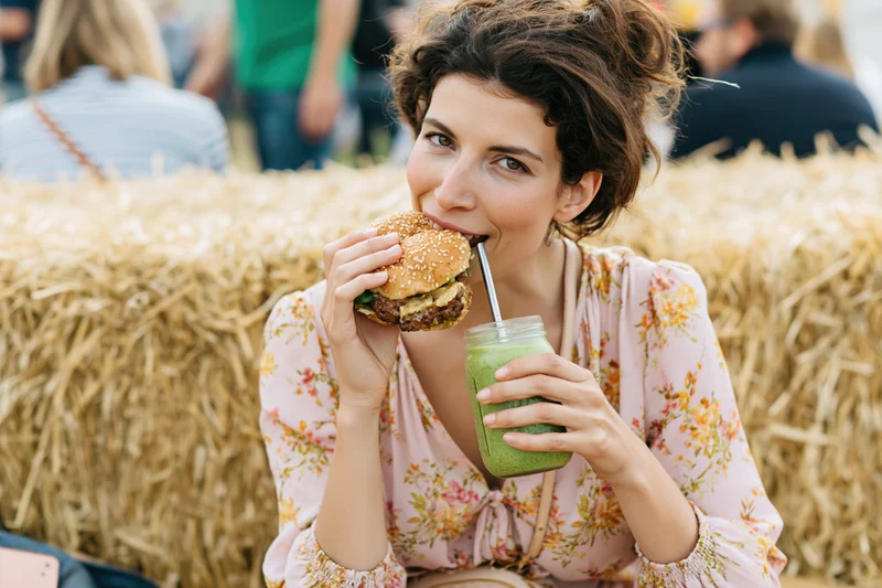 Woman biting into a burger while holding a green smoothie in a mason jar at an outdoor festival with hay bales behind her