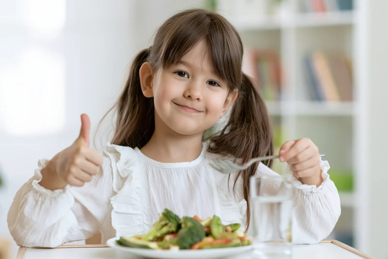 Young girl smiling and giving thumbs up while eating vegetables at a table