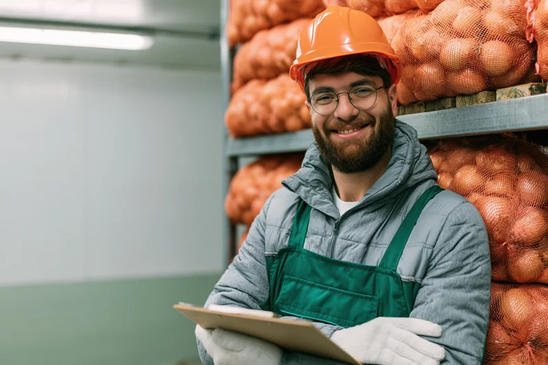 Young male warehouse worker smiling while holding clipboard and leaning against shelving stacked with net bags of onions in refrigerated storage