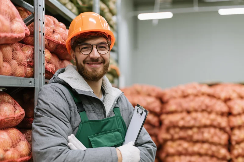 Young male warehouse worker smiling with clipboard leaning against a shelf of onions in a storage facility