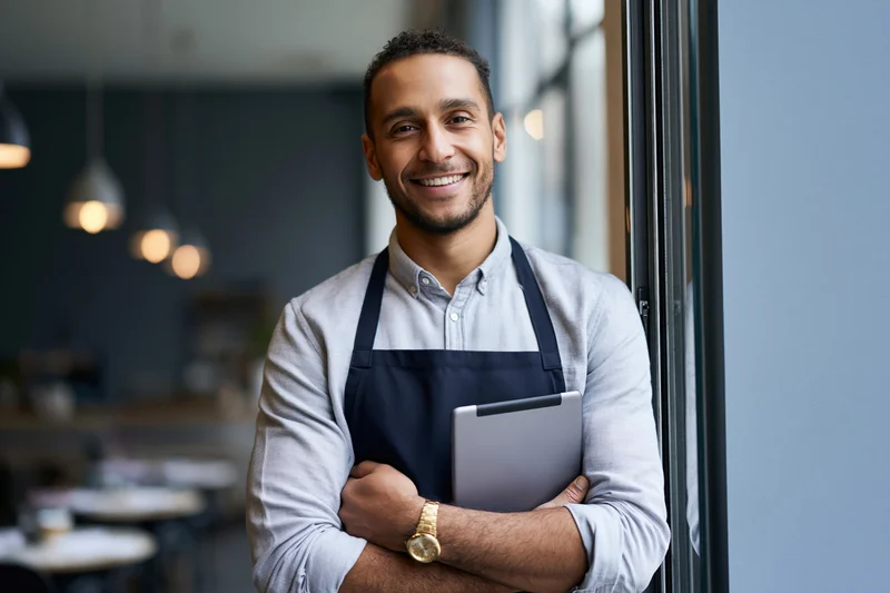 Smiling man in apron holding tablet while leaning against glass door in cafe interior