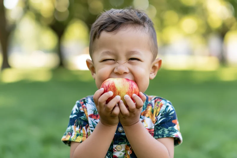 Young mixed-race toddler boy biting into a red apple in a grassy park