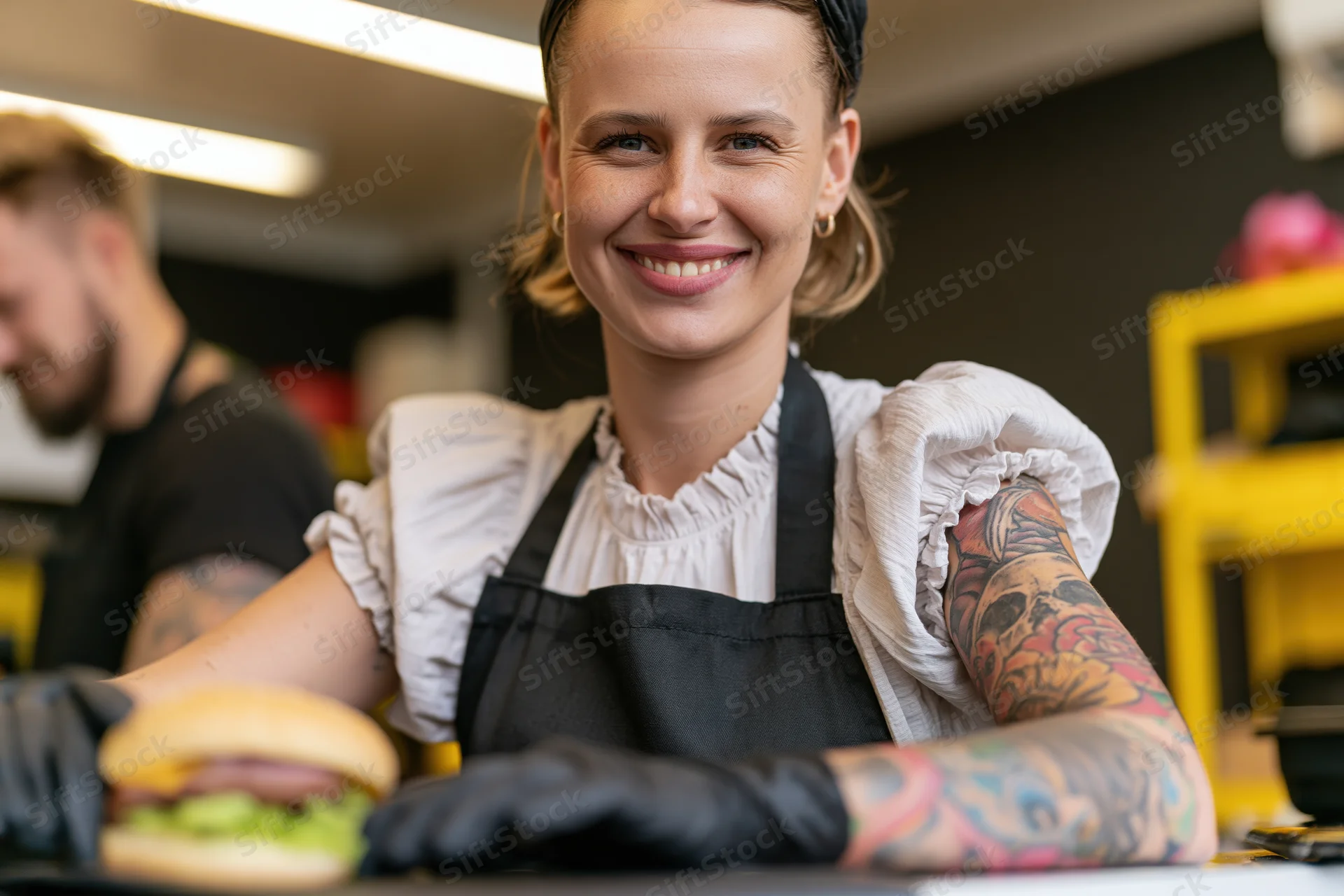 Smiling tattooed woman in apron and gloves holding burger in small food stall kitchen