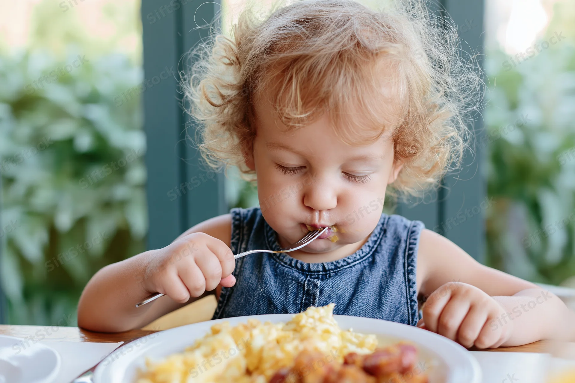 Toddler eating breakfast with fork at dining table near window