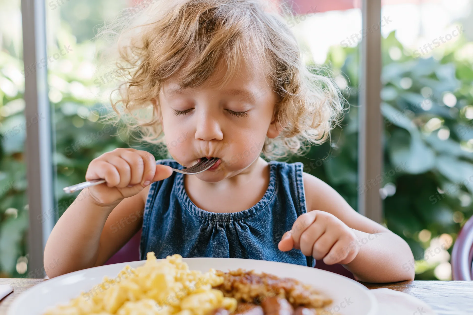 Toddler with curly blonde hair eating spaghetti and meat from a plate at a wooden table outdoors