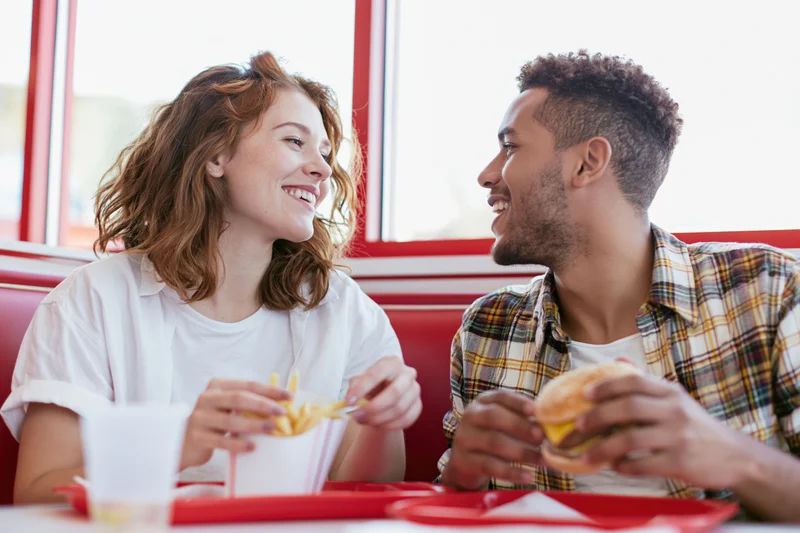 Young couple smiling and eating fast food together at a red booth in a diner