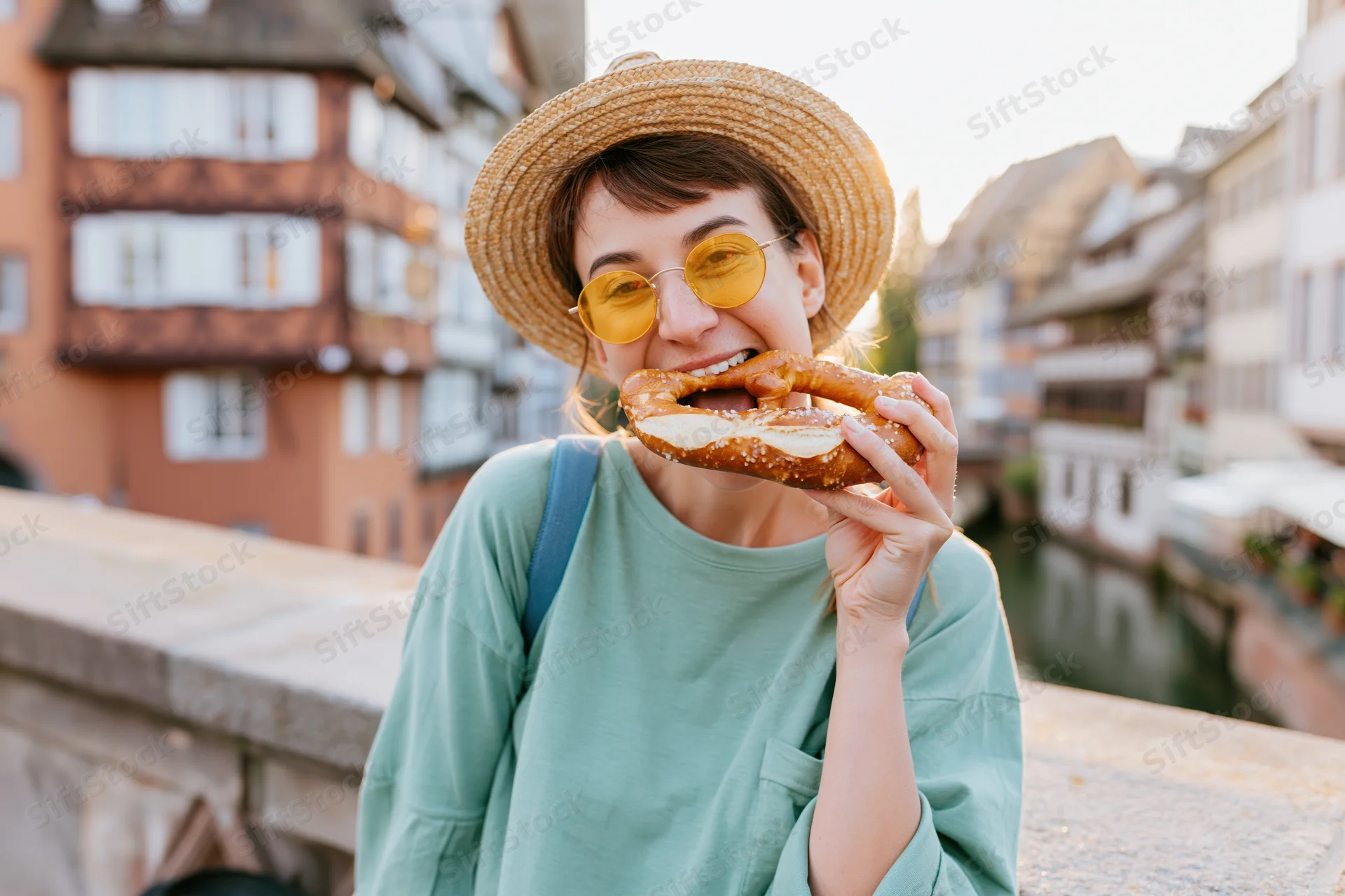 Woman eating a large soft pretzel on a stone bridge over a canal in a historic european city