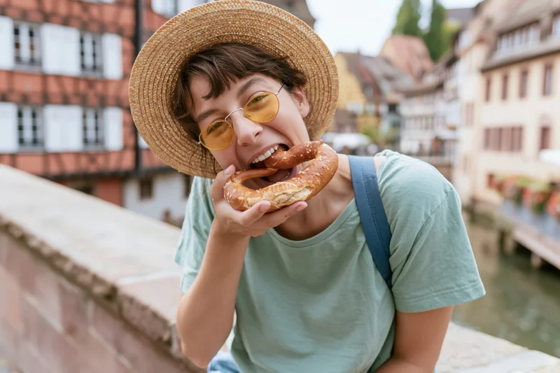 Woman eating a large soft pretzel on a stone bridge over a canal in a historic european town