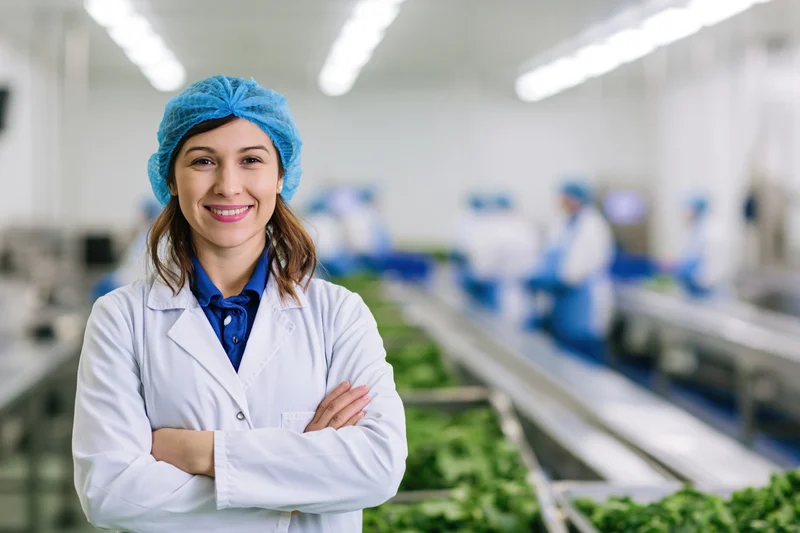 Woman in labcoat and hairnet standing with arms crossed in food factory production line