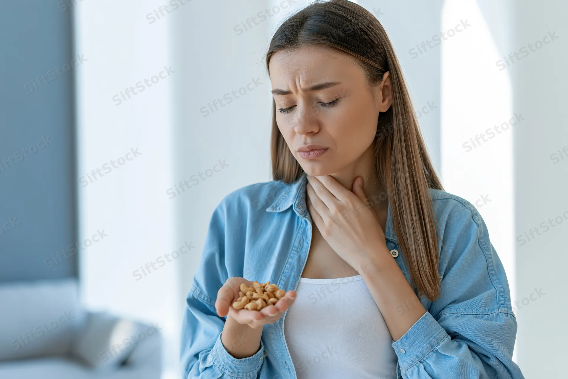 Young woman holding cashews and touching her throat in discomfort indoors