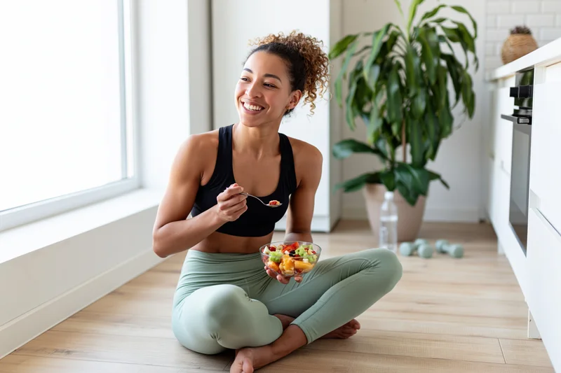 Woman sitting on kitchen floor eating fruit salad while smiling