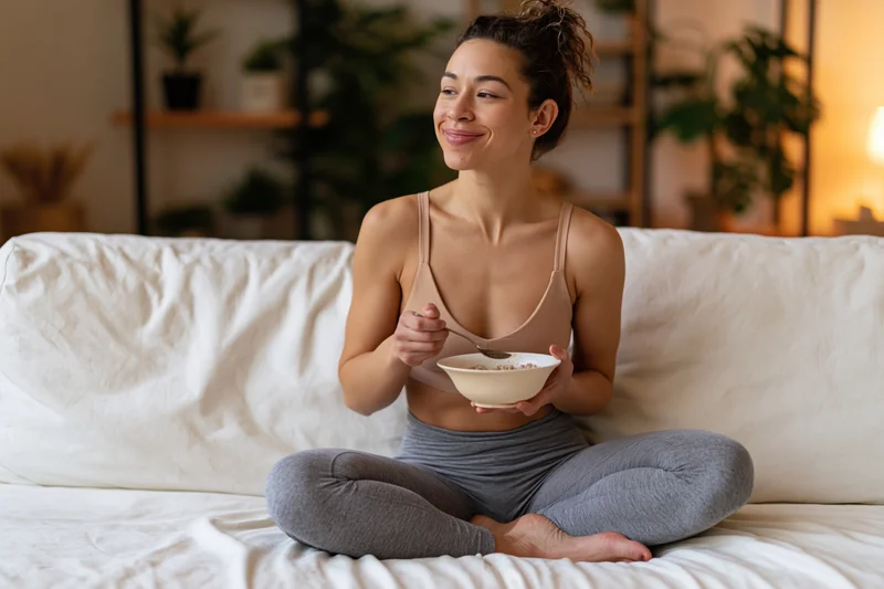 Woman sitting cross-legged on bed eating cereal from a bowl