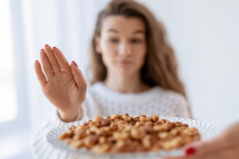 Woman pushing away a glass plate of mixed nuts with her hand in a bright indoor setting