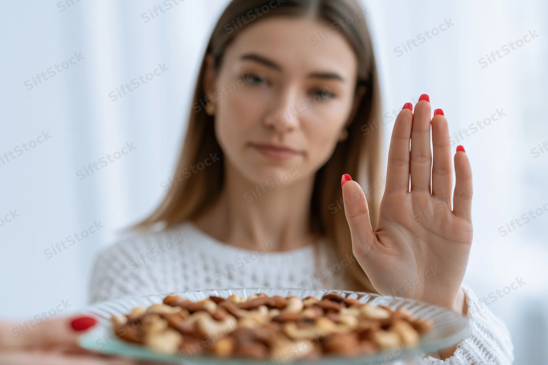 Woman pushing away a glass plate of mixed nuts with her hand in a bright indoor setting