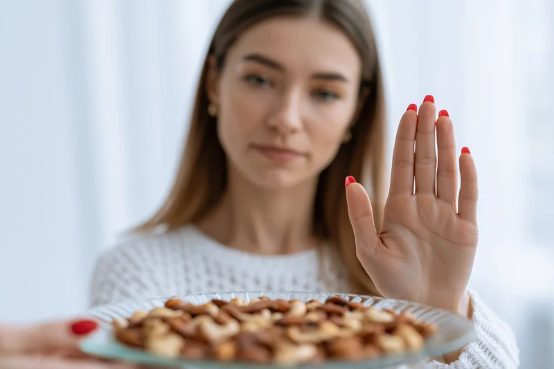 Woman pushing away a glass plate of mixed nuts with her hand in a bright indoor setting