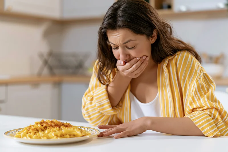 Woman covering her mouth in disgust at a plate of pasta on a kitchen table