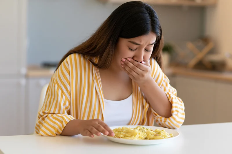 Woman covering her mouth in disgust while sitting at a kitchen table with a plate of pasta in front of her