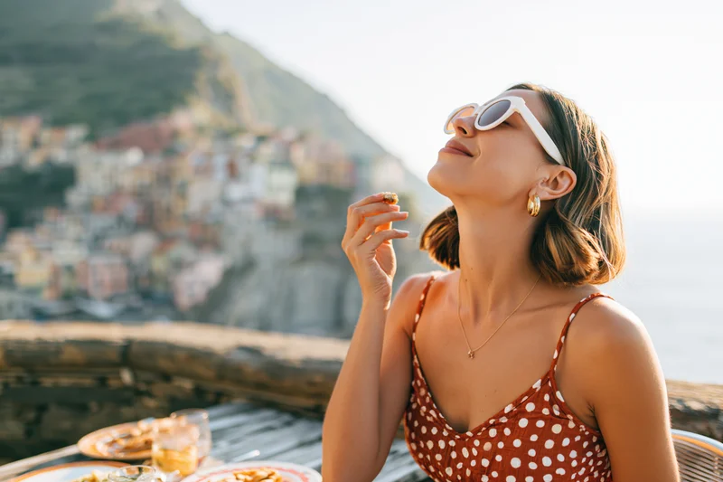 Woman smiling with sunglasses at outdoor terrace table overlooking coastal village and sea