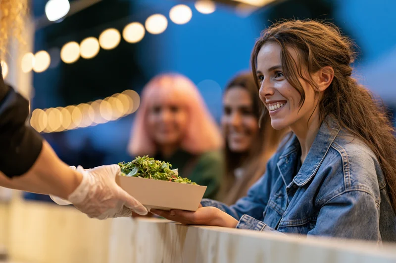 Young woman smiling as she receives a paper takeaway box of salad from a vendor at an outdoor evening event