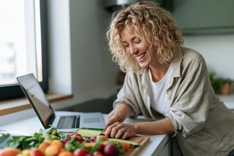 Woman smiling while chopping vegetables on a kitchen countertop next to an open laptop