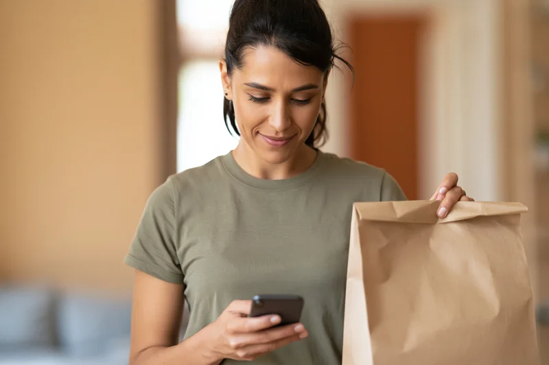 Woman holding paper bag and looking at smartphone at home