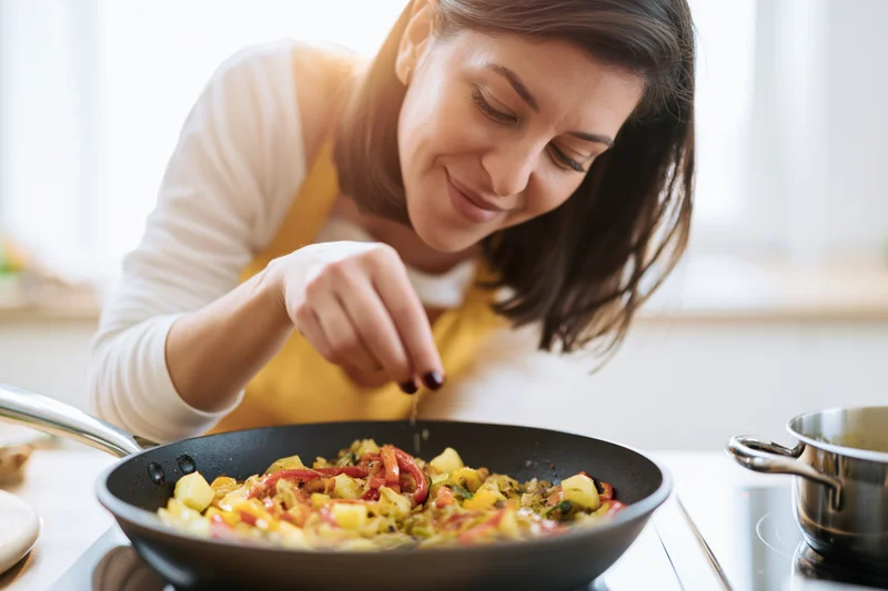 Woman smiling while seasoning stir fry in a kitchen pan on stove