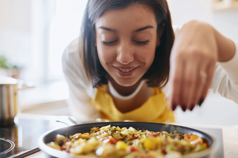 Young woman smiling while seasoning a colorful vegetable dish in a kitchen